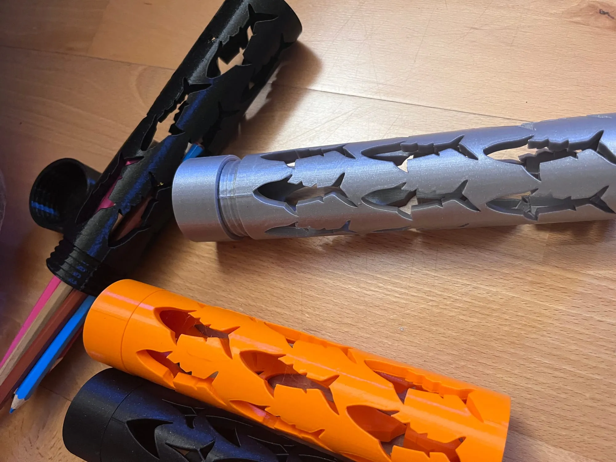 Étuis cylindriques ajourés argent, orange et noir posés sur une table en bois avec des crayons de couleur